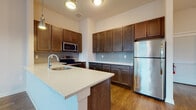 This thoughtfully designed single-floor unit offers a cohesive blend of modern finishes and functional living spaces. The kitchen serves as a standout feature, showcasing rich dark wood cabinetry extending to the ceiling for ample storage, light quartz countertops, a subway tile backsplash in a soft blue-gray tone, and a full suite of stainless steel appliances including a range, microwave, and refrigerator. A peninsula with a built-in sink and pendant lighting overhead creates an open, welcoming workspace ideal for cooking and entertaining. Warm hardwood-style flooring runs throughout the main living areas and bedrooms, lending a sense of continuity and warmth. The bedrooms are bright and airy, each featuring large windows that invite generous natural light and offer views of the surrounding neighborhood. A split-panel white interior door adds a clean, contemporary touch. The unit includes two full bathrooms: one featuring a glass-enclosed walk-in shower with subway tile surround and a dark wood vanity, and the other offering a full tub-shower combination with matching tile, a vanity, and light stone-look tile flooring. Together, these spaces create a cohesive, modern living environment with quality finishes throughout.