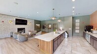Located at 550 S Windsor Ln in Flagstaff, Arizona, this thoughtfully designed single-floor community space blends comfort and sophistication across a well-appointed open layout. The living area serves as the heart of the space, anchored by a striking whitewashed brick fireplace wall adorned with brass sconce lighting, a wall-mounted television, and curated artwork, including a large framed landscape painting. Plush, rounded lounge chairs and a wooden coffee table create an inviting gathering spot atop warm area rugs set over light hardwood flooring that runs throughout. The kitchen and bar area features a long, white quartz-topped island lined with cognac leather and gold-toned bar stools, pendant lighting overhead, and a dedicated coffee bar station complete with appliances, dark cabinetry, and a display case. Sage green millwork, paneled walls, and coordinated doors lend the space a cohesive, elevated character. The dining area is framed by expansive floor-to-ceiling grid-pane windows that flood the room with natural light and offer sweeping views of a resort-style outdoor pool and lounge deck. The overall ambiance is warm yet polished, combining earthy tones, natural textures, and contemporary finishes to create a welcoming and functional communal environment.