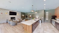 Located at 550 S Windsor Ln in Flagstaff, Arizona, this thoughtfully designed single-floor community space blends comfort and sophistication across a well-appointed open layout. The living area serves as the heart of the space, anchored by a striking whitewashed brick fireplace wall adorned with brass sconce lighting, a wall-mounted television, and curated artwork, including a large framed landscape painting. Plush, rounded lounge chairs and a wooden coffee table create an inviting gathering spot atop warm area rugs set over light hardwood flooring that runs throughout. The kitchen and bar area features a long, white quartz-topped island lined with cognac leather and gold-toned bar stools, pendant lighting overhead, and a dedicated coffee bar station complete with appliances, dark cabinetry, and a display case. Sage green millwork, paneled walls, and coordinated doors lend the space a cohesive, elevated character. The dining area is framed by expansive floor-to-ceiling grid-pane windows that flood the room with natural light and offer sweeping views of a resort-style outdoor pool and lounge deck. The overall ambiance is warm yet polished, combining earthy tones, natural textures, and contemporary finishes to create a welcoming and functional communal environment.