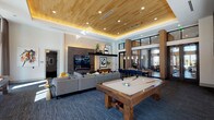 Located at 11576 Pearland Pkwy in Houston, TX, this single-story clubhouse amenity space is thoughtfully designed to offer residents a sophisticated and versatile gathering environment. The main lounge commands attention with soaring ceilings clad in warm wood paneling, ambient cove lighting, and a wall-mounted entertainment center set against a textured stone accent wall. A full-size billiards table anchors the room, complemented by a plush sectional seating area and a shuffleboard table positioned along floor-to-ceiling windows that flood the space with natural light and offer views of the outdoor surroundings. Adjacent to the main lounge, a dedicated media room provides an intimate retreat, featuring deep wraparound sectional sofas, blackout drapery, curated gallery wall art, and recessed cove lighting for a true screening-room atmosphere. The space transitions seamlessly into a well-appointed dining and kitchen area, highlighted by a striking granite-topped communal table beneath elegant pendant lighting, rich wood cabinetry, subway tile backsplash, and full kitchen appliances. The entry foyer offers a grand welcome with double glass doors, tall columns, and art-adorned walls, setting an upscale tone throughout the entire facility.