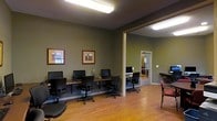 This single-floor space is designed as a practical and well-organized computer lab or business center, offering a comfortable environment for work and productivity. The main area features a row of desktop computer workstations lined along a shared desk surface against one wall, each equipped with a monitor, keyboard, and ergonomic office chair — providing ample capacity for multiple users simultaneously. The walls are painted in a muted olive green tone, lending the space a calm, professional atmosphere, and are accented with framed artwork that adds a touch of warmth. Warm-toned hardwood flooring runs throughout, contributing to an inviting yet functional aesthetic. Fluorescent ceiling fixtures provide consistent, even lighting across the space. An open doorway leads into an adjacent room, which contains an additional workstation and connects to a further area with a glass-paneled door, suggesting the space extends into supplementary utility or office rooms. A small seating area near the entrance, featuring a cushioned chair, offers a waiting or informal gathering spot. The overall layout is efficient and straightforward, making this space well-suited for shared computing, remote work, or administrative functions.