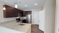 This single-floor space centers around a well-appointed kitchen that combines style and practicality. The kitchen showcases rich, dark espresso cabinetry that extends to the ceiling, providing generous storage throughout. Light granite countertops with subtle veining offer a striking contrast against the dark cabinetry, while a peninsula with an undermount sink creates a natural divide between the kitchen and adjacent living areas, offering additional prep and casual dining space.

Stainless steel appliances, including a French door refrigerator, electric range, and over-the-range microwave, lend a contemporary feel to the space. A pendant light fixture positioned above the peninsula adds focused ambient lighting, complementing the recessed ceiling lights that illuminate the room evenly.

The flooring throughout is a dark hardwood-style material that ties the overall aesthetic together cohesively. Neutral warm-toned walls keep the space feeling open and inviting. A white paneled door near the kitchen leads to an adjacent room, further highlighting the clean, crisp design details found throughout this thoughtfully designed space.
