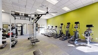 Located at 1801 Eureka Rd in Roseville, California, this fitness facility offers a thoughtfully organized exercise space designed to support a wide range of workout routines. The single-floor layout provides an open, spacious feel that accommodates multiple training zones without feeling cramped.

The cardio section is lined with a row of treadmills and elliptical machines positioned along a bold chartreuse accent wall, creating an energizing visual focal point. Overhead, a large ceiling fan and a grid of recessed panel lighting ensure the space remains comfortable and well-lit throughout. Natural light filters in through windows, further brightening the environment.

The strength training area is well-appointed, featuring a multi-functional cable and pulley system, a power rack, and a full dumbbell rack with a mounted flat-screen television above for entertainment during workouts. A large wall mirror spans one section, enhancing the sense of depth and allowing users to monitor their form.

The functional training zone includes stability balls, BOSU trainers, medicine balls, and battle ropes, offering versatile options for balance and conditioning work. The flooring throughout combines durable carpet tile with harder surface zones, delineating training areas while providing comfort underfoot. The overall design is clean, modern, and practical, making this facility a comprehensive destination for fitness in the Roseville community.