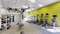 Located at 1801 Eureka Rd in Roseville, California, this fitness facility offers a thoughtfully organized exercise space designed to support a wide range of workout routines. The single-floor layout provides an open, spacious feel that accommodates multiple training zones without feeling cramped.

The cardio section is lined with a row of treadmills and elliptical machines positioned along a bold chartreuse accent wall, creating an energizing visual focal point. Overhead, a large ceiling fan and a grid of recessed panel lighting ensure the space remains comfortable and well-lit throughout. Natural light filters in through windows, further brightening the environment.

The strength training area is well-appointed, featuring a multi-functional cable and pulley system, a power rack, and a full dumbbell rack with a mounted flat-screen television above for entertainment during workouts. A large wall mirror spans one section, enhancing the sense of depth and allowing users to monitor their form.

The functional training zone includes stability balls, BOSU trainers, medicine balls, and battle ropes, offering versatile options for balance and conditioning work. The flooring throughout combines durable carpet tile with harder surface zones, delineating training areas while providing comfort underfoot. The overall design is clean, modern, and practical, making this facility a comprehensive destination for fitness in the Roseville community.