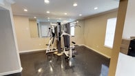 This fitness center is a thoughtfully designed, single-level facility offering a comprehensive workout environment. The space is organized across several connected open areas, allowing for clear separation between cardio and strength training zones. Polished dark flooring runs throughout, lending a sleek, professional aesthetic that is both durable and easy to maintain.

The strength training area features a Paramount multi-station cable machine positioned in front of a full-length wall mirror, which enhances the sense of space and allows users to monitor their form during exercise. The cardio zone is equipped with a True treadmill, a True elliptical trainer, and a True recumbent exercise bike, providing a well-rounded selection of aerobic options.

Natural light enters through windows fitted with vertical blinds, complementing the recessed ceiling lighting that ensures consistent illumination throughout. Crown molding details along the ceilings add a refined architectural touch. The facility also includes built-in shelving, a small storage or utility area, and fire safety equipment. A dedicated restroom is accessible directly from the main fitness space, contributing to the overall convenience and functionality of the center.