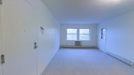 This single-floor space offers a clean, minimalist layout designed for straightforward functionality. The main room is finished with wall-to-wall carpeting in a neutral tone, complementing the crisp white walls and white ceiling that give the space a bright, airy feel. Two side-by-side windows allow natural light to fill the room, creating an open and welcoming atmosphere. Beneath the windows sits a wall-mounted air conditioning unit, providing climate control. A baseboard heating system runs along the perimeter, ensuring year-round comfort. A white paneled door with standard hardware is visible on one side of the room, contributing to the clean, uniform aesthetic. On the opposite wall, a small electrical panel or intercom fixture is mounted, indicating basic built-in utilities. The entry door features a deadbolt and handle lock for security. The overall design is simple and uncluttered, with no ornate architectural details, making it a versatile space adaptable to a variety of uses. The single-floor layout ensures ease of accessibility throughout.