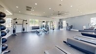 Located at 316 Columbia Dr in Carrollton, GA, this fitness center offers a generously sized, single-floor layout designed to accommodate a comprehensive workout experience. The main exercise area features durable rubber flooring and is outfitted with a strong selection of cardio equipment, including multiple treadmills and elliptical machines, all positioned beneath wall-mounted flat-screen TVs and a large ceiling fan that keeps the space comfortable. Large windows along the perimeter fill the room with natural light while offering views of the surrounding exterior. The strength training section features a dumbbell rack, weight benches, a cable machine, and a multi-functional strength station, with floor-length mirrors reflecting the open space and amplifying the sense of depth. A dedicated studio room branches off from the main floor, finished with light wood-style flooring and a cooler gray accent wall. This room is equipped with spin bikes, resistance bands, medicine balls, and wall-mounted storage — ideal for cycling classes or functional training. Recessed lighting and additional mirrors make it feel bright and open. The bathroom provides convenient access between the two areas, featuring tile flooring and a clean, functional layout to support post-workout needs.