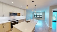 Located at The Breeze in Long Beach, NY, this thoughtfully designed 2-bedroom, 2-bathroom residence offers a modern open-concept layout with clean lines and contemporary finishes throughout. Light wood-tone flooring flows seamlessly across the entire living space, complementing the neutral warm-gray palette that creates a bright, cohesive ambiance.

The kitchen is a standout feature, boasting a large waterfall-edge island with an undermount sink and a professional-style spring-neck faucet. Two-toned cabinetry — combining white high-gloss upper cabinets with natural wood lower cabinets — pairs with crisp white quartz countertops and a full white backsplash. Stainless steel appliances, including a French door refrigerator, electric range, and built-in microwave, complete this chef-inspired space. Stylish Edison-bulb pendant lights hang above the island, connecting the kitchen to the open living area, which features large windows that invite abundant natural light and city views.

The spacious primary bedroom opens directly to a private balcony through floor-to-ceiling sliding glass doors, offering elevated outdoor views. Both bedrooms feature generous closets with wire shelving systems for organized storage. The bathrooms are finished with tile flooring, wood-toned vanities, and white quartz countertops. Two hallways connect the layout efficiently, rounding out this well-appointed residence.