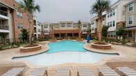 This amenity space is part of a well-established residential community characterized by classic brick and neutral-toned exterior architecture spanning multiple stories. The centerpiece of the outdoor area is a generously sized, freeform swimming pool with striking turquoise water, framed by a broad stamped concrete deck and raised brick planters. Tall palm trees and manicured landscaping border the pool area, adding a resort-like ambiance, while lounge chairs positioned along the deck invite relaxation.

The fitness center is accessed through a glass-paneled entry door and is organized across a well-lit, open layout with light-toned, patterned carpet tiles underfoot. Multiple ceiling fans are mounted throughout, ensuring comfortable air circulation during workouts. Recessed lighting and wall-mounted flat-screen televisions contribute to a functional and modern atmosphere. Natural light filters in through a set of multi-pane windows, offering views toward the surrounding building exterior.

Cardiovascular equipment, including multiple treadmills and stationary bikes, lines one section of the fitness area. A dedicated strength-training zone features a multi-function resistance machine alongside additional equipment. The neutral white walls and clean crown molding detailing throughout give the fitness center a polished, well-maintained appearance that complements the overall community aesthetic.