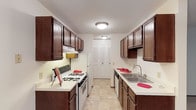 This single-floor residential space is defined by its efficiently designed galley-style kitchen, which serves as a central highlight. The kitchen features rich, dark wood cabinetry lining both sides of the corridor layout, providing generous storage through both upper and lower cabinets. White appliances, including a gas range with oven and a dishwasher, complement the light countertops and create a clean, classic contrast against the dark cabinetry. A stainless steel sink with a standard faucet is positioned along one wall, adding a practical focal point to the workspace. The flooring consists of neutral-toned tile with a subtle pattern, contributing to the bright and tidy feel of the kitchen. Overhead, flush-mount ceiling fixtures provide consistent, even lighting throughout the space. A textured ceiling adds a traditional touch to the interior. At the far end of the kitchen, a set of bifold doors suggests a closet or pantry, adding further storage utility. The overall layout is thoughtfully arranged to maximize functionality within a compact footprint, making excellent use of every inch of available space while maintaining a cohesive and orderly aesthetic throughout.