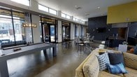 This single-floor communal lounge is designed with a contemporary aesthetic, combining industrial and modern elements throughout its open layout. Polished concrete floors anchor the space and reflect natural light streaming in through a striking row of clerestory windows positioned high along the upper walls, ensuring consistent brightness throughout the day. Large glass double doors along one wall open to an outdoor area, further enhancing the sense of openness and connectivity to the exterior.

The space is organized into distinct activity zones. A pool table positioned toward one side serves as a central recreational feature, complemented by industrial-style bar stools and high-top tables nearby for casual gathering. A comfortable lounge area with patterned upholstered sofas and accent pillows provides a relaxed setting for conversation or leisure.

On the opposing wall, a bold dark stone accent wall provides a dramatic architectural focal point, housing a wall-mounted television above a fireplace. Additional seating clusters with side tables are arranged nearby, creating an intimate atmosphere within the larger open plan. Subtle pendant lighting and recessed ceiling fixtures contribute to the overall warm, inviting ambiance of the space.