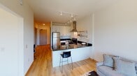 This well-appointed one-bedroom unit is designed with a clean, modern aesthetic throughout. The open-concept kitchen serves as a functional centerpiece, featuring stainless steel appliances — including a full-size refrigerator, range with a stainless steel hood vent, and dishwasher — paired with sleek dark granite countertops and crisp white cabinetry. A peninsula with bar seating connects the kitchen to the living area, making it an ideal space for casual dining or entertaining. Track lighting overhead ensures the kitchen is well-lit at all times. Warm hardwood flooring runs throughout the main living areas, adding a sense of continuity and warmth to the space. The bedroom is a tranquil retreat, offering ample natural light through a large window that frames views of the surrounding greenery and urban landscape. A wall-mounted HVAC unit provides individual climate control. The bathroom is finished with matching dark granite countertops, white cabinetry with brushed nickel hardware, and bright vanity lighting above a large mirror. An adjacent closet with built-in shelving and hanging space provides generous storage. The overall palette of soft whites and warm neutrals throughout creates a calm, cohesive, and inviting living environment.