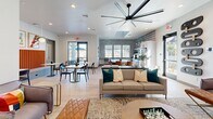 Located at 13215 W Glendale Ave in Glendale, Arizona, this single-floor community lounge is a thoughtfully designed gathering space that blends modern aesthetics with comfortable functionality. The space is anchored by a living area featuring a sleek linear fireplace set beneath a large wall-mounted television, creating a striking focal point. Stylish globe wall sconces provide warm ambient lighting, complementing the recessed ceiling lights and light wood-tone flooring found throughout. Plush accent chairs in muted tones, paired with leather ottomans and a round gold coffee table, are arranged over a textured area rug, encouraging relaxed conversation. Bold abstract artwork adds vibrant splashes of color to the otherwise crisp, light-gray walls. Adjacent to the lounge is a casual dining nook highlighted by a vivid orange upholstered banquette, marble-topped tables, and pendant lighting — all framed by framed graphic art prints. The main open area features a generous neutral sofa with teal accent pillows beneath a statement oversized ceiling fan, with access to an outdoor area through full-height glass doors that invite natural light deep into the space. A patterned geometric accent wall adds visual depth, while views into a fitness center are visible through a glass-paneled door, underscoring the space's role as a multifaceted community hub.