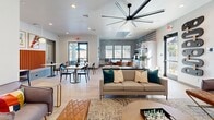 Located at 13215 W Glendale Ave in Glendale, Arizona, this single-floor community lounge is a thoughtfully designed gathering space that blends modern aesthetics with comfortable functionality. The space is anchored by a living area featuring a sleek linear fireplace set beneath a large wall-mounted television, creating a striking focal point. Stylish globe wall sconces provide warm ambient lighting, complementing the recessed ceiling lights and light wood-tone flooring found throughout. Plush accent chairs in muted tones, paired with leather ottomans and a round gold coffee table, are arranged over a textured area rug, encouraging relaxed conversation. Bold abstract artwork adds vibrant splashes of color to the otherwise crisp, light-gray walls. Adjacent to the lounge is a casual dining nook highlighted by a vivid orange upholstered banquette, marble-topped tables, and pendant lighting — all framed by framed graphic art prints. The main open area features a generous neutral sofa with teal accent pillows beneath a statement oversized ceiling fan, with access to an outdoor area through full-height glass doors that invite natural light deep into the space. A patterned geometric accent wall adds visual depth, while views into a fitness center are visible through a glass-paneled door, underscoring the space's role as a multifaceted community hub.