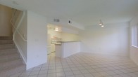 This two-floor space is designed with a clean, open-concept layout that emphasizes simplicity and functionality. The main living area is finished with large, light-toned ceramic tile flooring that extends throughout, creating a cohesive and easy-to-maintain surface underfoot. The white walls and ceiling contribute to a bright, airy atmosphere, while recessed HVAC vents are neatly integrated into the ceiling for a streamlined look. A track light fixture provides directional lighting to the living space.

A half-wall partition serves as a visual and functional divider between the living area and the kitchen, creating definition without sacrificing the open feel. The kitchen is equipped with white cabinetry, a range with a built-in overhead exhaust hood, and under-cabinet lighting that illuminates the workspace effectively. A countertop bar along the partition offers a casual dining or prep surface. The kitchen's slightly warmer lighting contrasts subtly with the cooler tones of the living space. Overall, the design prioritizes practicality and a neutral palette, making the space highly adaptable to a variety of furnishing styles.