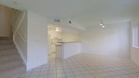 This two-floor space is designed with a clean, open-concept layout that emphasizes simplicity and functionality. The main living area is finished with large, light-toned ceramic tile flooring that extends throughout, creating a cohesive and easy-to-maintain surface underfoot. The white walls and ceiling contribute to a bright, airy atmosphere, while recessed HVAC vents are neatly integrated into the ceiling for a streamlined look. A track light fixture provides directional lighting to the living space.

A half-wall partition serves as a visual and functional divider between the living area and the kitchen, creating definition without sacrificing the open feel. The kitchen is equipped with white cabinetry, a range with a built-in overhead exhaust hood, and under-cabinet lighting that illuminates the workspace effectively. A countertop bar along the partition offers a casual dining or prep surface. The kitchen's slightly warmer lighting contrasts subtly with the cooler tones of the living space. Overall, the design prioritizes practicality and a neutral palette, making the space highly adaptable to a variety of furnishing styles.