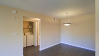 This single-floor space combines practical functionality with clean, understated design. The main room is finished with rich, dark hardwood flooring that contrasts cleanly against the white baseboards and neutral, warm-toned walls, creating a bright and welcoming atmosphere. A stylish pendant light fixture hangs from the ceiling, adding a subtle decorative focal point while providing overhead illumination to the space.

A dedicated in-unit laundry room is accessible directly from the main area through a wide doorway. This laundry space is equipped with a full-size washer and dryer and features built-in shelving above the units, offering convenient storage for laundry essentials and household items. The laundry room also includes an additional door, suggesting further access to adjacent storage or utility areas.

The overall layout is straightforward and efficient, with a neutral color palette throughout that lends itself well to a variety of furnishing styles. The combination of hardwood flooring, in-unit laundry, and ample wall space makes this a highly functional and adaptable living environment.