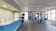 This fitness center offers a thoughtfully designed workout environment spread across a single floor. The space features a clean, modern aesthetic with two-tone walls in soft gray and bold blue, complemented by recessed lighting and ceiling fans that ensure proper ventilation and comfort during workouts. Large windows allow natural light to fill the space, creating an energizing atmosphere. The main workout area is anchored by a multi-station cable and weight machine, ideal for a full range of strength training exercises. A dedicated free weights zone includes a medicine ball rack stocked with various sizes, offering versatility for functional fitness routines. The flooring transitions between patterned carpet tiles and durable rubber matting, clearly delineating workout zones and providing appropriate traction and cushioning. Full-length mirrors line one wall, allowing users to monitor form and technique. A built-in counter with a light stone surface near the entrance adds a practical element, suitable for checking in or storing personal items. The overall layout is open and spacious, allowing for comfortable movement between equipment stations and accommodating a variety of fitness activities.