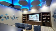 This single-floor media lounge is a thoughtfully designed entertainment space that blends modern aesthetics with functional comfort. The room's most striking feature is its ceiling design, where large circular acoustic panels are dramatically backlit with vibrant blue LED lighting, creating a cinematic ambiance that immediately sets the mood upon entry. The focal point of the room is a large-format display screen mounted within a built-in dark wood entertainment wall, flanked by open shelving on both sides — providing both visual appeal and practical storage. The seating arrangement features plush, velvet-upholstered chairs and sofas in a cool grey-blue tone, organized to provide clear sightlines to the screen from every angle. Low-profile coffee tables are positioned throughout the seating area for added convenience. Light wood-look flooring grounds the space with warmth, offering a pleasing contrast to the cool tones of the lighting and upholstery. Film reel decorations adorn one wall, reinforcing the cinematic theme of the space. Recessed lighting complements the LED accents, ensuring the room is well-lit while preserving its immersive atmosphere. Overall, this space is ideal for screenings, presentations, or social gatherings.