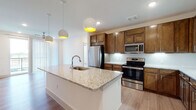 This single-floor residence showcases a thoughtfully designed open layout that seamlessly connects the kitchen and living areas. The kitchen serves as a central highlight, featuring rich medium-toned wood cabinetry extending to the ceiling for generous storage, light granite countertops, and a full suite of stainless steel appliances including a French door refrigerator, range, and built-in microwave. A large center island with a built-in sink anchors the space, illuminated by a trio of modern globe pendant lights with warm yellow interiors. A white penny-tile backsplash adds texture and contrast against the warm cabinetry. Light wood-tone flooring runs throughout the main living areas, contributing to the bright, airy feel of the space.

The living area opens directly to a private balcony through sliding glass doors dressed with vertical blinds, allowing ample natural light. A ceiling fan provides comfort overhead. The bedroom is carpeted and features a dedicated window with an exterior view and its own ceiling fan. The ensuite bathroom offers dual granite-topped vanities with wood cabinetry, a glass-enclosed walk-in shower, and direct access to a spacious walk-in closet outfitted with multiple shelving tiers and hanging rods.