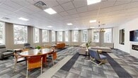 This thoughtfully designed single-floor amenity space offers an impressive and versatile environment built for both productivity and relaxation. The centerpiece is a generously sized open lounge area wrapped in a curved wall of large windows, flooding the space with abundant natural light and offering views of the surrounding landscape. Patterned carpet tiles in muted blues, grays, and golds define the lounge zones, which are furnished with comfortable sectional sofas, accent chairs, and round tables suited for casual gatherings or informal meetings. A refined chandelier adds an elegant touch overhead. Adjacent to the lounge, a fully equipped kitchen features stainless steel appliances, a multi-seat island with bar stools, and a striking floor-to-ceiling coffee bean mural, creating a vibrant and inviting break area with colorful pendant lighting. The fitness room, visible through interior windows, provides a dedicated workout space. A game area with a ping pong table offers recreational appeal. Private rooms with light wood flooring, wall-mounted displays, and flexible seating arrangements serve as focused meeting or collaboration spaces. Blueprint-style artwork displayed throughout adds a cohesive, professional aesthetic to this dynamic, multi-use facility.