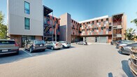 Located at 163 Artful Way in Asheville, North Carolina, this outdoor parking facility is situated directly alongside a striking contemporary multi-story building characterized by a bold facade of warm brick tones, metal paneling, and a grid of windows and balconies. The building's modern architectural design features exposed steel stairwells, industrial-style lighting, and a mix of dark brick and corrugated metal cladding that gives the complex a distinctive urban character.

The parking lot itself is thoughtfully laid out with clearly marked stalls and directional arrows guiding traffic flow. The lot wraps around portions of the building and is accessible from multiple points, providing convenient access. A concrete retaining wall borders one side of the lot, while mature trees and lush greenery provide natural screening and shade along the perimeter, softening the overall environment.

The surrounding streetscape reflects Asheville's vibrant and growing urban character, with neighboring multi-story buildings visible in the distance. The site benefits from open sky exposure and abundant natural light throughout the day. Bike racks are also present near building entry points, supporting alternative transportation options. Overall, this well-maintained surface lot offers practical, accessible parking in a dynamic and architecturally engaging Asheville setting.