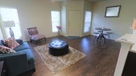 This single-floor residence offers a welcoming and functional layout centered around an open living area. The space is anchored by rich, dark hardwood-style flooring that extends throughout, providing a warm and cohesive aesthetic against the neutral beige walls.

The living area is generously proportioned, accommodating a central seating arrangement complete with a tufted round leather ottoman resting atop a plush shag area rug, complemented by a patterned accent chair. A small bistro-style table with industrial bar stools is positioned near the entry, offering a casual dining or social nook.

The front entry is framed by a classic paneled door flanked by two windows that allow natural light to filter into the space, creating a bright and airy atmosphere. A decorative vase with floral accents adds a touch of warmth near the entry wall.

The overall ambiance is casual yet put-together, with the dark flooring serving as a strong visual foundation. The open arrangement of the living space promotes easy movement and adaptability, making it well-suited for both everyday living and casual entertaining. The single-floor layout ensures convenient accessibility throughout.