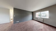 Located at 2311 Florida Dr in Fort Wayne, Indiana, this one-bedroom, one-bathroom unit offers a practical and functional layout across a single floor. The combined living and dining area is generously proportioned, featuring plush carpeting, a bold slate-gray accent wall, and a large window that allows natural light to fill the space. A wall-mounted radiator unit provides heating beneath the window, which offers views of the surrounding neighborhood.

The bedroom is a cozy, carpeted room with its own window and radiator, maintaining a consistent aesthetic with neutral wall tones. Three closets distributed throughout the unit provide ample storage for residents.

The kitchen features blue-gray cabinetry with a double stainless steel sink, light countertops, and wood-look flooring, with a window above the sink bringing in additional natural light. The bathroom includes classic tile wainscoting in cream and burgundy tones, a built-in storage cabinet, and a clean, functional layout.

Throughout the unit, exposed pipe columns add an industrial character, while consistent carpeting and neutral tones create a cohesive, understated ambiance. Multiple windows ensure natural light reaches nearly every room, making the space feel open and connected to its surroundings.