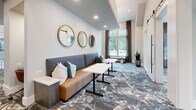 Located at 2307 Ridgemont Ave in Charlotte, NC, this thoughtfully designed single-floor amenity space offers a well-appointed layout comprising a living room and two distinct dining areas. The space immediately impresses with soaring ceilings and floor-to-ceiling windows that flood the interiors with natural light and provide open views of the surrounding neighborhood greenery.

The living area features a comfortable banquette-style sofa in warm cognac leather, accented with teal and white throw pillows. A trio of gold-framed circular mirrors anchors the wall above, while patterned carpeting in muted earth tones and blues adds visual depth and texture underfoot. Small café-style tables complement the seating arrangement, creating an inviting, lounge-like atmosphere.

The two dining areas are refined and functional. One is highlighted by dark accent walls, a bold geometric pendant light fixture, and a sleek white marble-top conference table surrounded by leather chairs, ideal for meetings or private gatherings. The other dining nook features the same marble-top table paired with vibrant abstract wall art in teal and gold, lending an artistic energy to the space.

Throughout, the design balances modern sophistication with approachable warmth, combining curated artwork, elegant lighting, and cohesive furnishings to create a versatile and welcoming communal environment.