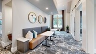 Located at 2307 Ridgemont Ave in Charlotte, NC, this thoughtfully designed single-floor amenity space offers a well-appointed layout comprising a living room and two distinct dining areas. The space immediately impresses with soaring ceilings and floor-to-ceiling windows that flood the interiors with natural light and provide open views of the surrounding neighborhood greenery.

The living area features a comfortable banquette-style sofa in warm cognac leather, accented with teal and white throw pillows. A trio of gold-framed circular mirrors anchors the wall above, while patterned carpeting in muted earth tones and blues adds visual depth and texture underfoot. Small café-style tables complement the seating arrangement, creating an inviting, lounge-like atmosphere.

The two dining areas are refined and functional. One is highlighted by dark accent walls, a bold geometric pendant light fixture, and a sleek white marble-top conference table surrounded by leather chairs, ideal for meetings or private gatherings. The other dining nook features the same marble-top table paired with vibrant abstract wall art in teal and gold, lending an artistic energy to the space.

Throughout, the design balances modern sophistication with approachable warmth, combining curated artwork, elegant lighting, and cohesive furnishings to create a versatile and welcoming communal environment.