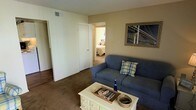 This single-story space presents a straightforward and functional layout designed for comfortable everyday living. The living room serves as the central gathering area, featuring carpeted flooring that adds warmth underfoot and neutral-toned walls that create a calm, understated ambiance. A blue sofa anchors the seating area, complemented by a white wooden coffee table that provides both storage and surface space. Wall art depicting a bridge adds a decorative focal point to the room.

An overhead HVAC vent is visible in the ceiling, indicating a central air system throughout the unit. The living area flows openly toward a hallway, which leads to at least one bedroom, visible through an open doorway. The bedroom appears carpeted as well, maintaining a consistent feel across the space. A closet door is also visible along the hallway, suggesting practical built-in storage.

The overall design is modest and utilitarian, with clean lines and simple finishes. The neutral color palette throughout the living area and connecting spaces contributes to a cohesive, livable environment that is easy to personalize.