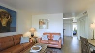 This single-floor residence offers a comfortable and well-appointed living environment with a thoughtfully designed open layout. The living area serves as the heart of the home, furnished with warm cognac leather sofas accented by gray throw pillows, centered around a rustic wood coffee table and a patterned area rug that adds texture underfoot. Wood-look flooring runs throughout, lending warmth and continuity to the space. A decorative mirror mounted on a dividing wall reflects the adjacent kitchen, offering a glimpse of wooden cabinetry and adding visual depth to the room. A classic table lamp with a fabric shade provides soft ambient lighting, complementing the natural light that filters in from the surrounding areas. The open floor plan allows for an easy flow between the living area and the rest of the home, with a hallway visible in the background leading to additional rooms. Clean white walls and smooth ceilings create a bright, airy atmosphere throughout. The overall ambiance is relaxed and inviting, combining functional furnishings with a neutral, earthy color palette that gives the space a grounded, livable character.
