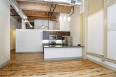 This single-floor unit showcases a distinctive loft-style design that blends industrial character with modern finishes. The open living area is defined by original hardwood floors with natural patina, soaring ceilings with exposed wooden beams, and visible ductwork that reinforces the space's industrial aesthetic. A ceiling fan with pendant lighting adds both function and character overhead.

Large windows fitted with horizontal blinds line the main living area, allowing abundant natural light to fill the space while offering views of the surrounding streetscape. Baseboard heating runs beneath the windows, ensuring year-round comfort.

The kitchen features dark espresso cabinetry extending to the ceiling for ample storage, complemented by richly veined granite countertops. A peninsula with a built-in sink and matching granite surface provides additional workspace and a casual dining option. Stainless steel appliances, including a refrigerator, range, and microwave, complete the well-equipped cooking area.

The bathroom is finished with gray tile walls, a sliding glass door shower enclosure, hardwood flooring, a white countertop vanity with dark cabinetry, and a large mirror, creating a clean and contemporary feel throughout.