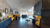 This sophisticated single-floor communal space showcases a striking blend of contemporary and industrial design elements. The main lounge area features bold yellow tufted banquette seating set against a textured exposed brick accent wall, complemented by dark large-format tile flooring and statement pendant lighting in brass and gold tones. A wall-mounted flat-screen TV adds a functional touch to this inviting gathering area. Dramatic vertical slat partitions divide the space while maintaining an open, airy feel, and floor-to-ceiling windows along the facade flood the interior with natural light and offer views of the surrounding landscape. A sleek dark credenza serves as a reception or display surface near the entry. The space transitions seamlessly into a games area anchored by a full-size billiards table set atop a richly patterned area rug, surrounded by eclectic vintage-inspired armchairs on decorative tile flooring. A polished concrete accent wall adds an industrial contrast to the warm brick. Beyond the games area, a glass-walled fitness center houses cardio equipment, maintaining visual connectivity throughout. The overall ambiance is upscale yet approachable, with layered textures, curated furnishings, and thoughtful lighting creating a dynamic and welcoming environment.