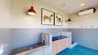 This dedicated pet grooming space in Huntsville, Alabama, is a compact yet fully equipped facility designed with both function and style in mind. The room features striking vibrant blue hexagonal floor tiles that immediately draw the eye, complemented by large-format gray wall tiles that extend halfway up the walls, creating a clean, two-tone aesthetic. A built-in terrazzo-trimmed wash station with a handheld sprayer provides a dedicated bathing area for pets, while a separate grooming counter with a dark surface and light wood cabinetry offers ample preparation space. Floor drains are strategically placed for efficient water drainage. Red pendant lights hang from the ceiling, adding a bold design accent and warm illumination throughout the room. A wall-mounted split HVAC unit ensures year-round climate comfort. Large floor-to-ceiling windows along one wall flood the space with natural light and provide views of the surrounding outdoor greenery. A glass-paneled entry door offers easy access. Playful framed artwork depicting a dachshund and a French bulldog adorns the walls, reinforcing the space's pet-friendly character. Leash and accessory hooks are mounted conveniently within reach, rounding out this well-appointed grooming station.