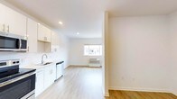 This single-floor space offers a seamless open-plan layout connecting the kitchen and living area, creating a functional and airy environment. The kitchen is outfitted with white cabinetry, a stainless steel sink, a dishwasher, and light countertops, all contributing to a clean, contemporary look. Recessed lighting in the kitchen ensures the workspace is well-illuminated. Light hardwood-style flooring runs throughout the entire unit, adding warmth and continuity between the two zones. The living area benefits from a centrally positioned window that allows natural light to filter in, enhancing the bright, open feel of the space. A wall-mounted HVAC unit beneath the window provides climate control. The walls are finished in a crisp white, complementing the neutral palette and reinforcing the minimalist character of the space. Structural columns subtly define the transition between the kitchen and living area without disrupting the overall sense of openness. The space presents a practical, well-designed layout suitable for everyday living, with an emphasis on clean lines and a light, welcoming atmosphere throughout.