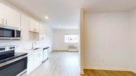 This single-floor space offers a seamless open-plan layout connecting the kitchen and living area, creating a functional and airy environment. The kitchen is outfitted with white cabinetry, a stainless steel sink, a dishwasher, and light countertops, all contributing to a clean, contemporary look. Recessed lighting in the kitchen ensures the workspace is well-illuminated. Light hardwood-style flooring runs throughout the entire unit, adding warmth and continuity between the two zones. The living area benefits from a centrally positioned window that allows natural light to filter in, enhancing the bright, open feel of the space. A wall-mounted HVAC unit beneath the window provides climate control. The walls are finished in a crisp white, complementing the neutral palette and reinforcing the minimalist character of the space. Structural columns subtly define the transition between the kitchen and living area without disrupting the overall sense of openness. The space presents a practical, well-designed layout suitable for everyday living, with an emphasis on clean lines and a light, welcoming atmosphere throughout.