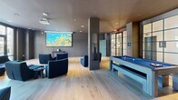 Located at 80 Batterson Park Rd in Farmington, CT, this contemporary amenity lounge delivers a well-appointed, single-floor entertainment experience. The open-plan layout is unified by wide-plank light hardwood flooring and a refined palette of deep grays and charcoal tones, creating a sophisticated yet inviting atmosphere throughout.

A striking focal point of the space is an artfully designed entertainment wall, adorned with an eclectic arrangement of audio speakers, vinyl records, and multimedia displays surrounding a central television — a bold, music-inspired installation that anchors one side of the room. Adjacent to this feature, a full-size billiards table with a blue felt surface provides a central recreational element.

A dedicated media area occupies another zone of the lounge, outfitted with a ceiling-mounted projector and a large pull-down screen, complemented by plush navy blue lounge chairs arranged for comfortable viewing. Structural columns divide the space subtly without interrupting its open flow.

Floor-to-ceiling steel-framed glass doors and windows flood the interior with natural light and offer direct views of and access to an outdoor patio area, seamlessly connecting the indoor and outdoor environments. A separate seating area features a leather sofa, coffee table, and additional accent chairs, ideal for relaxed conversation. Recessed ceiling lighting throughout ensures a consistently warm and well-lit ambiance.