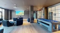 Located at 80 Batterson Park Rd in Farmington, CT, this contemporary amenity lounge delivers a well-appointed, single-floor entertainment experience. The open-plan layout is unified by wide-plank light hardwood flooring and a refined palette of deep grays and charcoal tones, creating a sophisticated yet inviting atmosphere throughout.

A striking focal point of the space is an artfully designed entertainment wall, adorned with an eclectic arrangement of audio speakers, vinyl records, and multimedia displays surrounding a central television — a bold, music-inspired installation that anchors one side of the room. Adjacent to this feature, a full-size billiards table with a blue felt surface provides a central recreational element.

A dedicated media area occupies another zone of the lounge, outfitted with a ceiling-mounted projector and a large pull-down screen, complemented by plush navy blue lounge chairs arranged for comfortable viewing. Structural columns divide the space subtly without interrupting its open flow.

Floor-to-ceiling steel-framed glass doors and windows flood the interior with natural light and offer direct views of and access to an outdoor patio area, seamlessly connecting the indoor and outdoor environments. A separate seating area features a leather sofa, coffee table, and additional accent chairs, ideal for relaxed conversation. Recessed ceiling lighting throughout ensures a consistently warm and well-lit ambiance.
