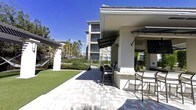This single-level outdoor amenity space is designed for community gathering and al fresco entertaining. The centerpiece is a covered outdoor kitchen and grilling station, complete with bar-height seating and a ceiling fan overhead to provide comfort in warm weather. A retractable awning offers additional shade over the cooking and dining area, making it a practical and inviting spot for residents year-round. The space is finished with attractive paver stonework that extends across the patio, creating a clean and polished aesthetic underfoot. Adjacent to the patio, a neatly manicured lawn provides open green space, bordered by decorative iron fencing that defines the area while maintaining an open, airy feel. Lush landscaping, including tropical palms and ornamental plantings, frames the space and adds a resort-like atmosphere. The surrounding multi-story residential buildings are visible in the background, reinforcing the communal nature of the amenity. Under clear, sunny skies, the entire area is bathed in natural light, enhancing the bright and welcoming ambiance. This thoughtfully designed outdoor space offers a seamless blend of functionality and comfort for everyday outdoor living.