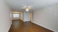 This single-floor residence presents a clean, well-maintained interior defined by rich, dark hardwood-style flooring that runs throughout the open living areas, creating a warm and cohesive aesthetic. The main living space is generously proportioned, offering ample room for a variety of furniture arrangements. A ceiling fan with an integrated light fixture provides both practical air circulation and ambient lighting. Built-in shelving units are seamlessly integrated into the wall, offering attractive and functional storage display space without occupying additional floor area. The open layout connects the living area to an adjoining room or dining space, which is illuminated by a decorative pendant chandelier and benefits from natural light through a window fitted with blinds. White interior doors and trim provide a crisp contrast against the neutral, warm beige walls, lending the space a bright and airy feel. A textured popcorn ceiling adds a classic residential character throughout. The layout also includes interior doors leading to additional rooms or storage, suggesting a thoughtfully organized floor plan designed for comfortable day-to-day living. Overall, the space conveys a practical yet inviting atmosphere suited for a range of lifestyles.