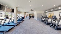 Located at 1083 E Sain St in Fayetteville, Arkansas, this dedicated fitness center offers a generously sized, single-floor exercise space designed to support a comprehensive workout experience. The layout is thoughtfully organized into distinct zones, maximizing functionality throughout the room.

The cardio area is expansive, housing a row of TRUE-brand treadmills, upright stationary bikes, and elliptical machines. Large windows and glass doors line the perimeter of this section, flooding the space with natural light and offering views of the surrounding outdoor environment. Ceiling fans are mounted overhead to promote airflow and comfort during use.

The strength training area is equally well-equipped, featuring multiple TRUE cable and selectorized weight machines, a dumbbell rack with a full range of free weights, adjustable benches, and a rowing machine. Wall-mounted flat-screen televisions are positioned throughout for entertainment during workouts.

The flooring throughout consists of durable, patterned carpet tiles in a neutral blue-gray tone, providing both cushioning and a clean aesthetic. Recessed lighting supplements the natural light, ensuring the space remains bright and inviting at all times. Decorative artwork accents the entryway area, adding a polished touch to the functional environment. Staircase access is also present within the space, connecting to adjacent building areas.