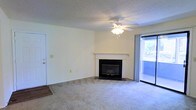 This single-floor space centers around a well-proportioned living area finished with wall-to-wall carpeting that adds warmth underfoot. A white-mantled fireplace with a dark surround serves as the room's focal point, providing both a functional heating element and a classic architectural detail. A ceiling fan with an integrated light fixture is centrally mounted on the textured ceiling, offering year-round climate comfort and ambient lighting. Natural light enters generously through a sliding glass door along one wall, which opens to an outdoor balcony or patio area, extending the living space and providing outdoor views. The walls are painted in a neutral, light tone that enhances the sense of openness throughout the room. A secondary door is visible along the adjacent wall, suggesting convenient access to additional areas of the unit. The overall layout is open and straightforward, with a clean, functional design that offers a versatile canvas for furnishing and personalization. The combination of the fireplace, natural light, and outdoor access gives the living area a comfortable and welcoming ambiance.