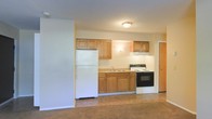 This single-floor space offers a straightforward, functional layout centered around an open kitchen and living area. The kitchen is equipped with warm-toned wooden cabinetry, both upper and lower, providing ample storage. Essential appliances are included: a full-size white refrigerator, a white electric range with a black oven, and a double-basin sink positioned beneath upper cabinets. The cabinetry and countertops are finished in neutral tones that complement the overall simplicity of the space. Flush ceiling lighting illuminates the kitchen area effectively. The adjoining living space features a open, unobstructed floor plan with cork-style flooring in a warm brown tone, offering a versatile area that can accommodate various furniture arrangements. The walls throughout are painted in a clean, neutral off-white, contributing to a bright and understated ambiance. A doorway visible near the kitchen provides access to an additional room or storage area. The overall design is practical and unpretentious, making efficient use of the available space with a clean, move-in-ready aesthetic.