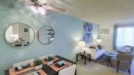 This single-floor space showcases an open-plan design that integrates the dining and living areas into one cohesive, functional layout. The living area is furnished with a white sofa accented by decorative throw pillows, complemented by a sleek black coffee table and a patterned area rug, creating a balanced, modern aesthetic. A pair of table lamps provides warm ambient lighting throughout the space.

The dining area features a dark-toned table set with place settings, decorative greenery, and coordinating chairs, offering a welcoming space for meals. The soft blue accent wall serves as a unifying backdrop across both areas, lending the space a calm and airy atmosphere.

Decorative round mirrors of varying sizes are mounted on the blue wall, adding visual depth and a stylish focal point. A colorful multi-panel artwork hangs in the living area, contributing a vibrant touch. The ceiling features a ceiling fan with an integrated light fixture, as well as a textured finish and a subtle beam detail. Carpet flooring runs throughout, adding warmth and comfort underfoot.