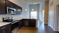 This single-floor space showcases a thoughtfully designed open-concept kitchen that immediately draws attention with its dramatically high ceilings, creating an airy, expansive atmosphere. The kitchen is anchored by sleek, dark espresso cabinetry that contrasts beautifully against light-toned granite countertops, offering generous prep and storage space. A peninsula with an undermount sink serves as both a functional workspace and a natural divider between the kitchen and the adjoining living area.

Stainless steel appliances, including a range, built-in microwave, and dishwasher, provide a polished, contemporary feel throughout. Pendant lighting hangs elegantly from the soaring ceiling above the peninsula, complementing the ceiling fan that helps circulate air throughout the open space. Rich, dark hardwood flooring extends across the entire area, grounding the design with warmth and continuity.

Natural light filters in through a window positioned at the rear of the space, brightening the interior and highlighting the clean lines of the cabinetry. A hallway visible along the right side of the space suggests additional rooms beyond, with a bright doorway enhancing the sense of openness and flow throughout this well-appointed home.
