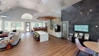 Located at 12825 Vinings Creek Dr in Matthews, NC, this single-floor community clubhouse delivers a polished, modern environment designed for both social gathering and productive work. The space is anchored by soaring vaulted ceilings that lend an expansive, airy atmosphere throughout. Rich hardwood-style flooring runs continuously across the open floor plan, tying together the distinct functional zones seamlessly.

At the heart of the space sits a striking central island topped with a dramatic marble-look surface, surrounded by sage green upholstered seating — ideal for casual meetings or communal gatherings. Adjacent to the island, a café-style service counter is equipped with beverage stations and white cabinetry, accented by a textured tile backsplash. A bold dark slate feature wall provides a strong visual counterpoint to the otherwise light and neutral palette.

A dedicated lounge area offers comfortable mixed seating arranged around a wall-mounted television and fireplace, framed by a decorative wood shelving unit set against patterned wallcovering. Large windows bathe this area in natural light and provide views of the surrounding outdoor greenery.

A glass-paneled sliding barn door leads to a private office or leasing suite, accented with colorful artwork and a herringbone-patterned floor. A grand arched window near the entry further enhances the architectural character of the space, completing this well-appointed and welcoming community hub.