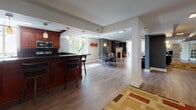 Located at 1450 Washington Blvd in Stamford, Connecticut, this well-appointed single-floor amenity space offers a diverse range of functional areas designed for entertainment and relaxation. The open-concept layout connects a fully equipped kitchen — outfitted with rich wood cabinetry, dark granite countertops, pendant lighting, and a breakfast bar with seating — to a bright dining area bathed in natural light from large windows. Adjacent to the dining space, a living area with a classic fireplace provides a warm, inviting atmosphere. The home theater room features a large wall-mounted flat-screen TV, built-in speakers, plush reclining leather seating, and patterned carpeting, creating an immersive cinematic experience, complemented by a retro popcorn machine for added charm. A dedicated game room houses a poker table surrounded by leather chairs, ideal for social gatherings. The lounge area showcases hardwood-style flooring, vibrant accent furniture, and curated wall art that adds pops of color throughout. The space also includes a billiards area, visible in the dollhouse view, and benefits from consistent recessed and sconce lighting throughout. Clean gray walls, white trim, and thoughtful transitions between areas give the entire space a cohesive, modern aesthetic.