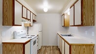 This single-floor, 1-bedroom, 1-bathroom unit is designed for efficient, comfortable living. The galley-style kitchen is well-appointed with ample wood-trimmed cabinetry on both sides, a white electric range, a full-size refrigerator, and a single-basin sink with generous counter space. Globe pendant lighting illuminates the kitchen, which features durable wood-look plank flooring that carries throughout the main living areas. The kitchen flows directly into the combined living room, which benefits from a large window that brings in natural light and offers outdoor views of surrounding greenery. A wall-mounted air conditioning unit provides climate control in the living area. The bedroom is a comfortable, well-proportioned retreat featuring soft carpeting, a ceiling fan with light fixture, a window with views of outdoor foliage, and a built-in double sliding-door closet for storage. The full bathroom is accessed via a hallway and includes a vanity with wood-trimmed cabinetry, a toilet, a bathtub, a towel bar, and matching wood-look flooring. A hallway connects the main living spaces and provides additional storage access. Neutral wall tones and consistent flooring throughout create a cohesive, clean aesthetic.