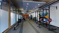 This fitness facility offers a thoughtfully designed workout environment that balances industrial aesthetics with modern functionality. The space features an exposed ceiling with visible ductwork and ventilation systems, complemented by a suspended panel with recessed lighting that illuminates the workout floor evenly. Large glass panels line one side of the space, allowing natural light to filter in and creating an open, airy atmosphere.

The main workout floor is finished with durable rubber flooring, featuring defined lane strips that add both style and functional utility for training drills. Equipment is organized efficiently throughout the space. A dedicated strength training section houses a power rack stocked with a full assortment of bumper plates in various weights, alongside medicine ball storage racks filled with colorful balls for functional fitness. A comprehensive dumbbell rack and adjustable benches are positioned for easy access.

Cardio and cable machines are arranged along the perimeter, while an orange accent wall at the far end adds a bold, energizing pop of color that enhances the overall motivating atmosphere. The generous ceiling height contributes to a spacious, non-restrictive feel throughout. The layout ensures ample room to move between stations, making this facility well-suited for both individual training sessions and group workout programming.